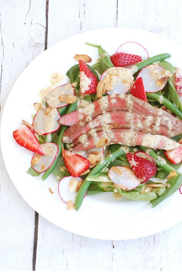 Umami-rich grilled flank steak tops a bed of baby spinach, crisp green beans, and sweet strawberries in this light summer steak salad drizzled with a sparkling, tangy Champagne shallot vinaigrette. via JessicaLevinson.com | #steaksaladrecipe #summersaladrecipe Summer Steak Salad Recipe on White Plate